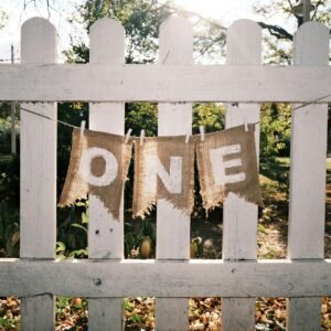 Outdoor scene of a burlap banner spelling 'ONE' on a white picket fence in autumn.